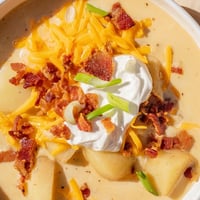 A steaming bowl of Loaded Baked Potato Soup topped with crispy bacon crumbles, sharp cheddar, and green onions on a rustic table.